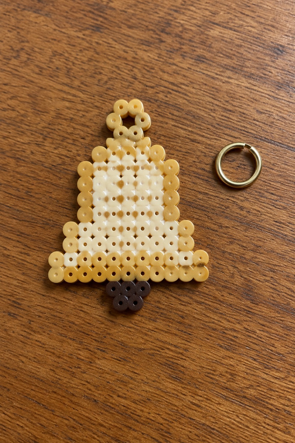 Yellow and white perler bead bell ornament with gold jump ring on wood.