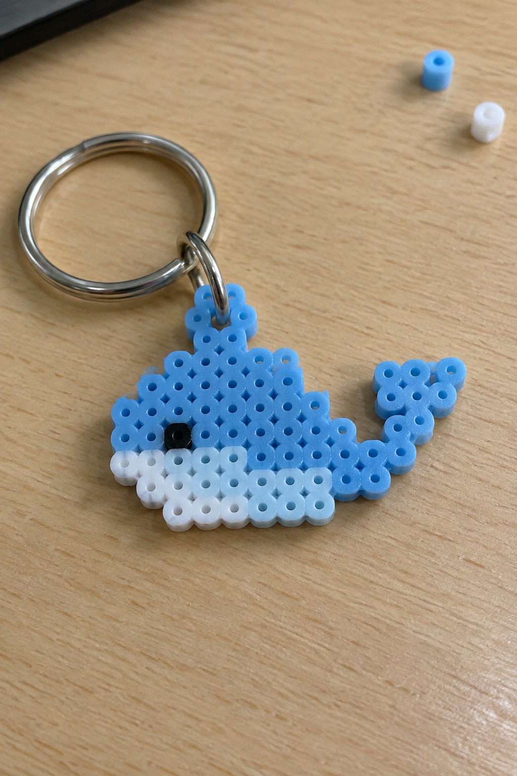 Blue and white perler bead whale keychain on wooden table with loose beads