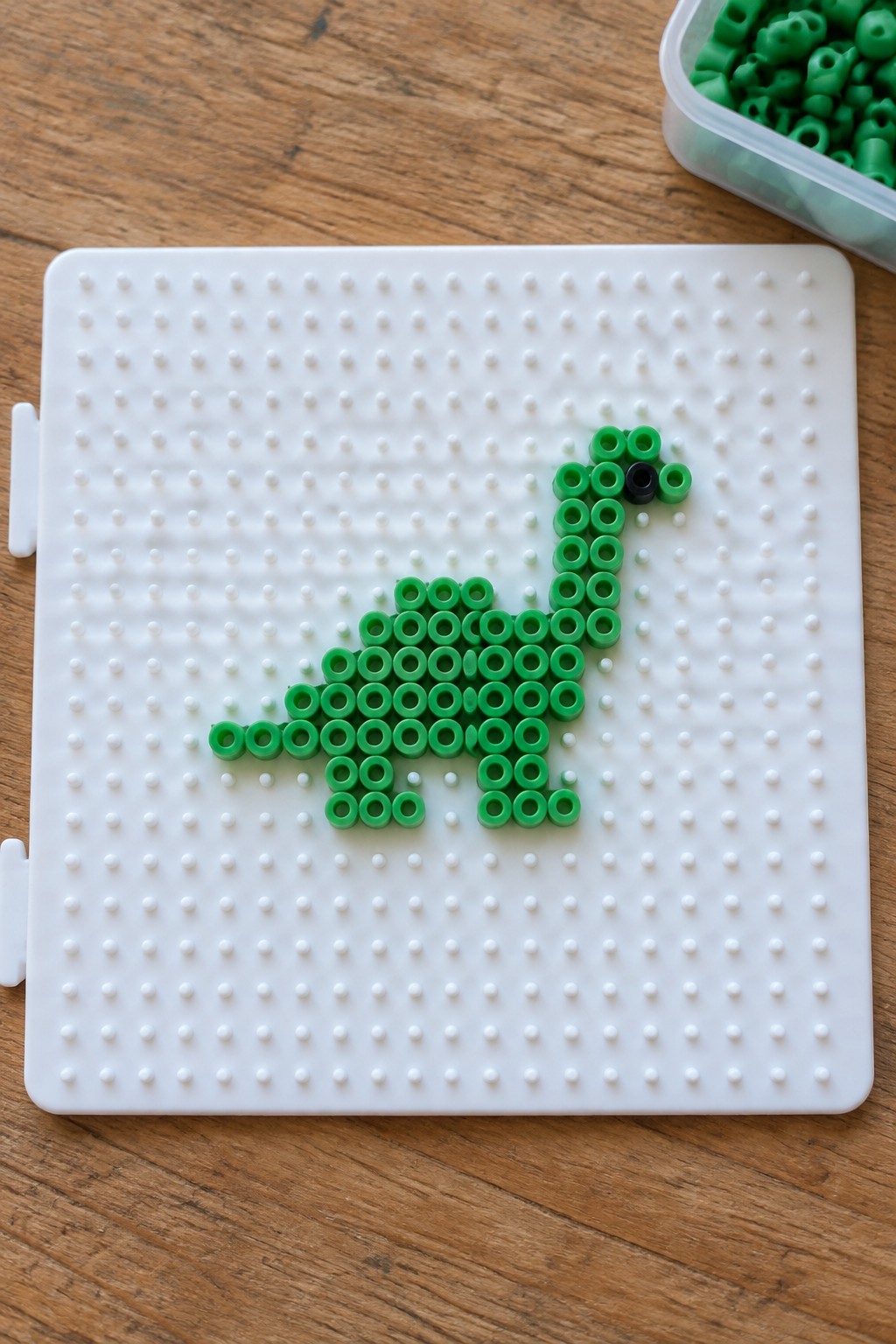 Green Perler bead dinosaur with black eye on a white pegboard.