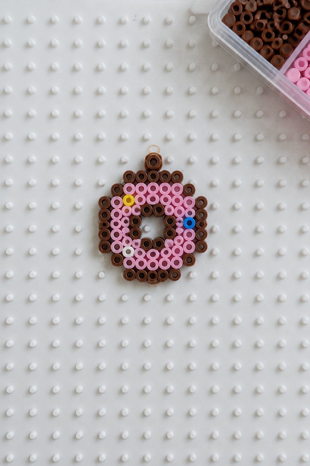 Perler bead donut pattern with brown base, pink icing, and colorful sprinkles on a white pegboard.