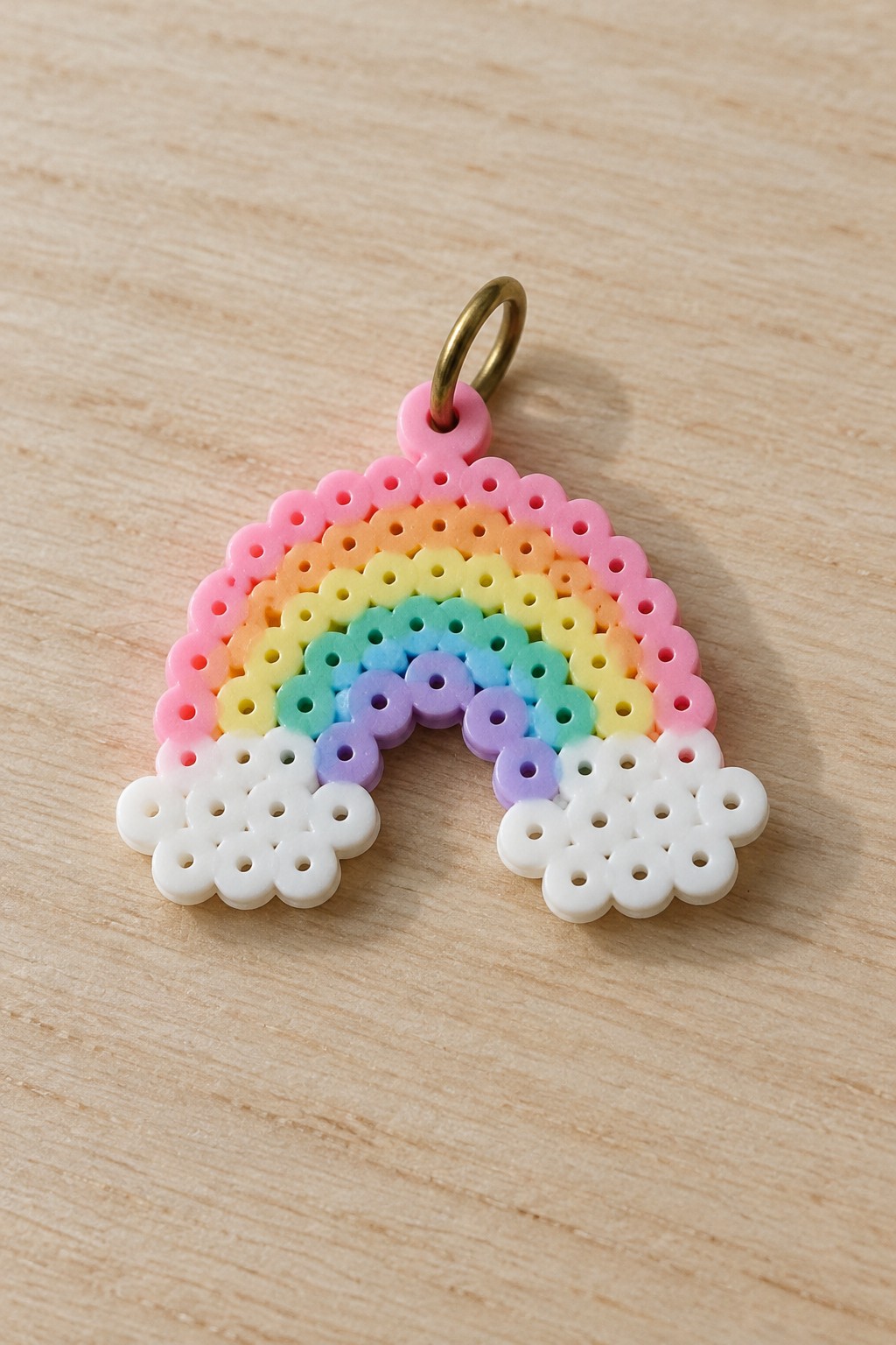 Rainbow perler bead charm with colorful arcs, white clouds, and gold ring on wood