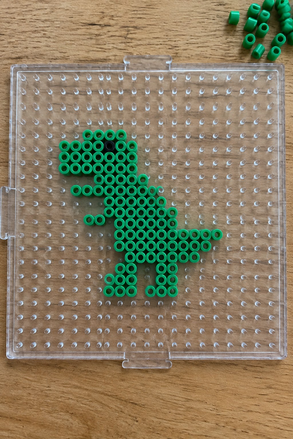 Green perler beads forming a T-rex with black eye on clear pegboard.