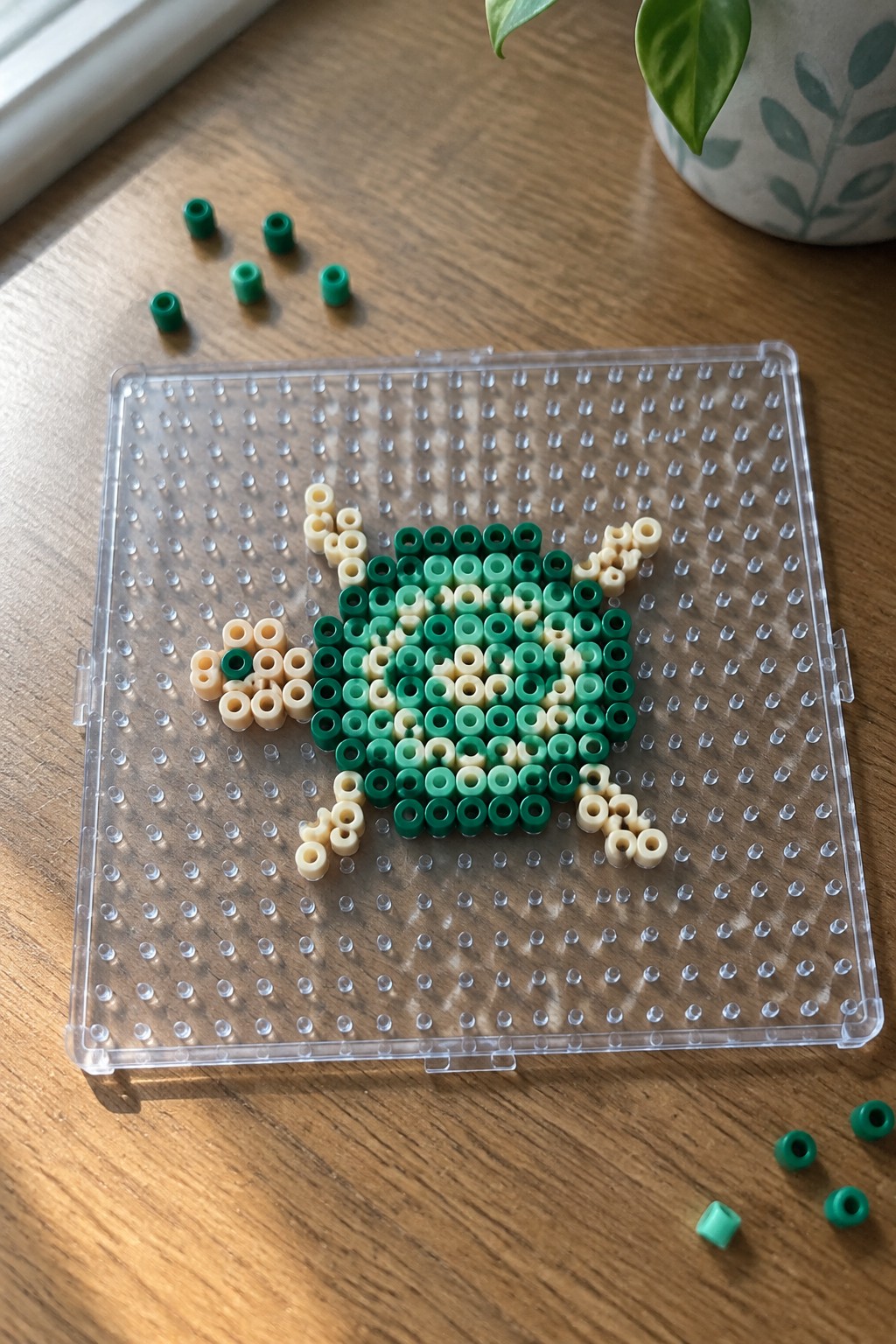 Green and beige perler beads forming a turtle on a clear pegboard with loose beads nearby