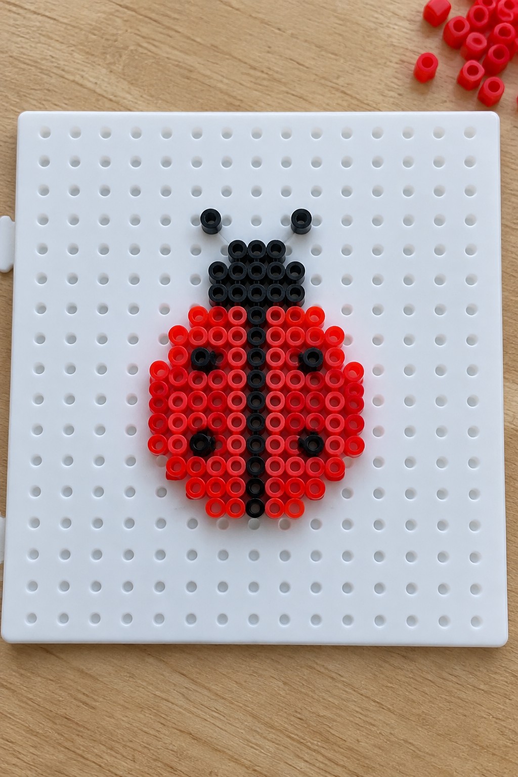 Red and black perler bead ladybug on white pegboard with loose beads nearby.