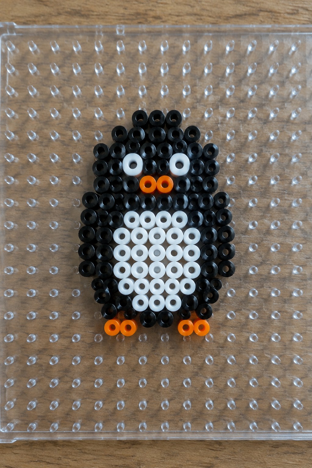 A small penguin made from black, white, and orange Perler beads arranged on a clear pegboard.