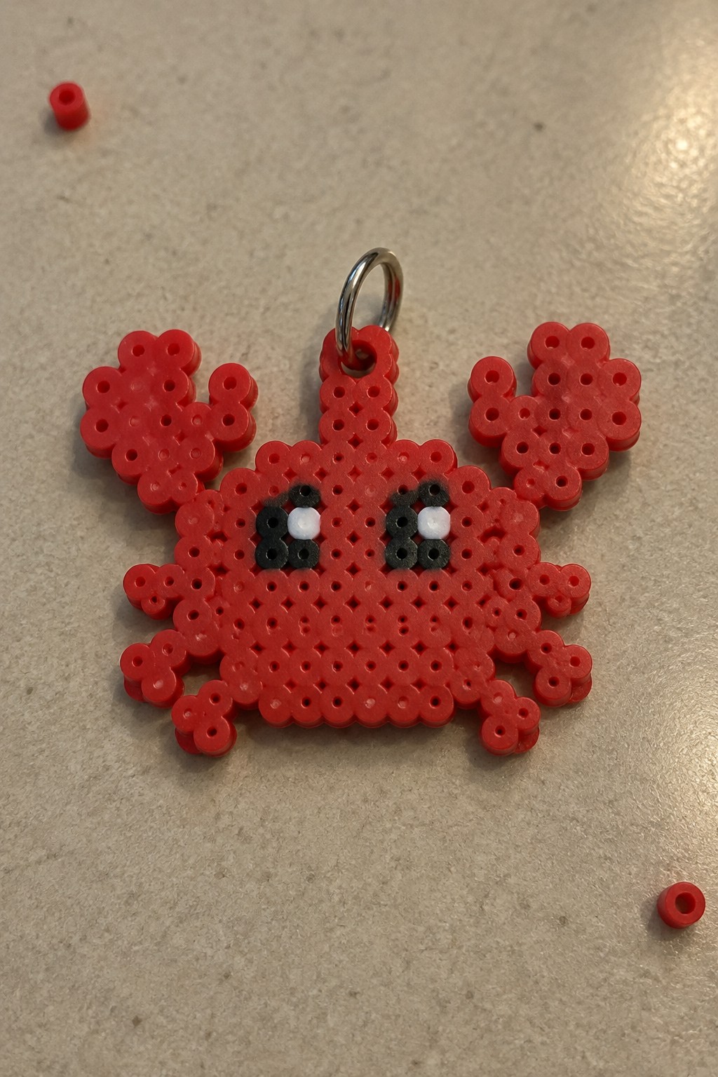 Red Perler bead crab with black and white eyes on a pegboard, metal ring attached at the top.