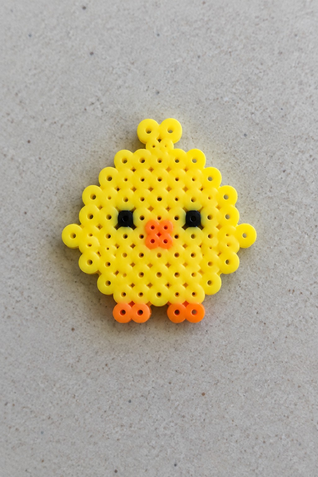 Unfused Perler bead chick on a pegboard with yellow body, black eyes, orange beak and feet, and a hanging loop at the top.