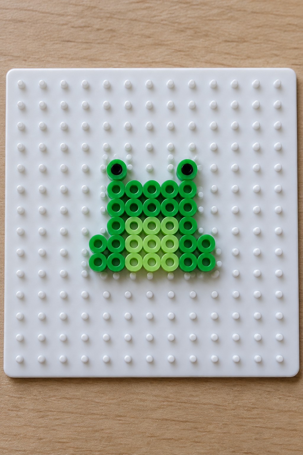 A small frog made with green and black Perler beads arranged on a white pegboard.