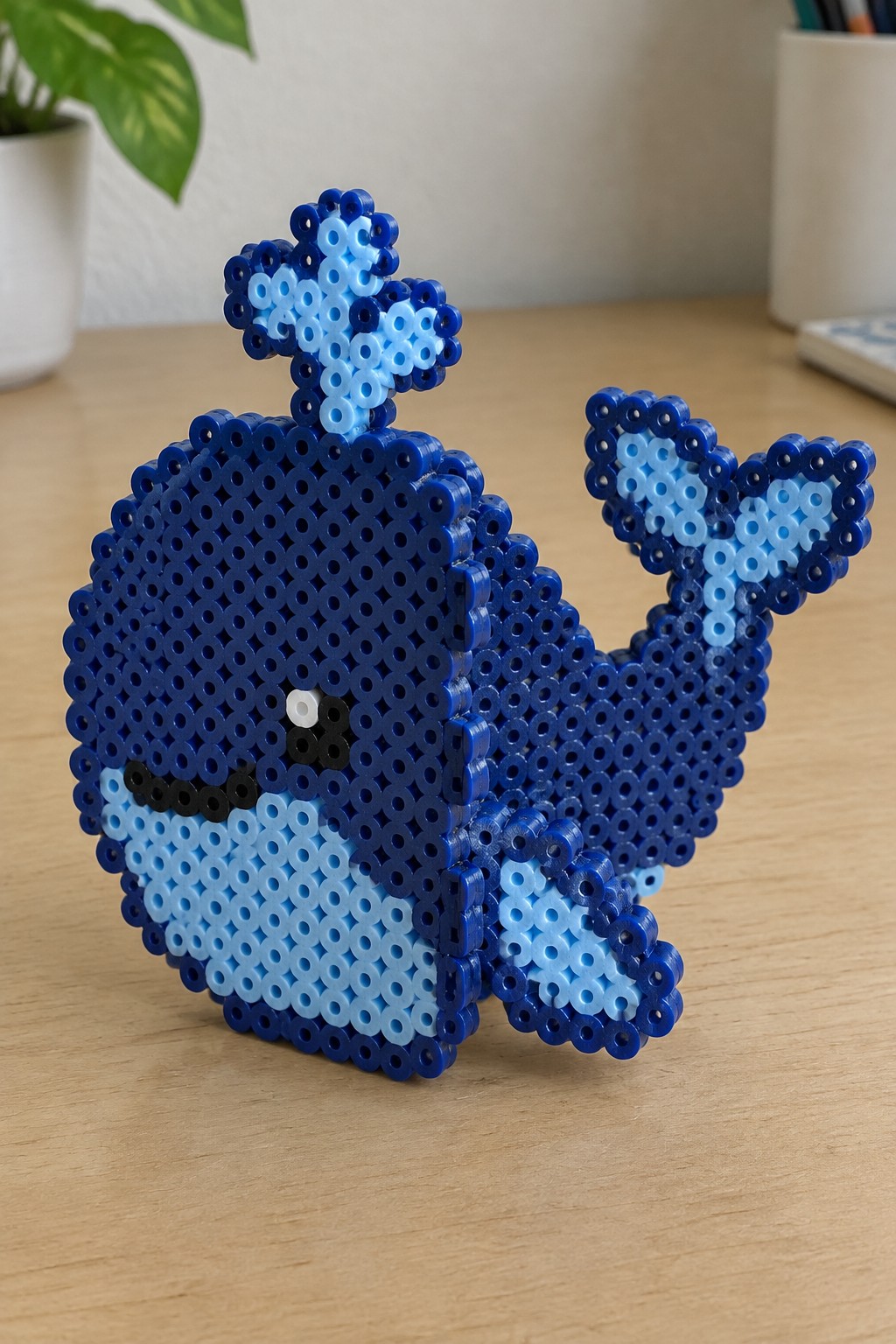 Blue perler bead whale with light blue belly, spout, and tail on wood