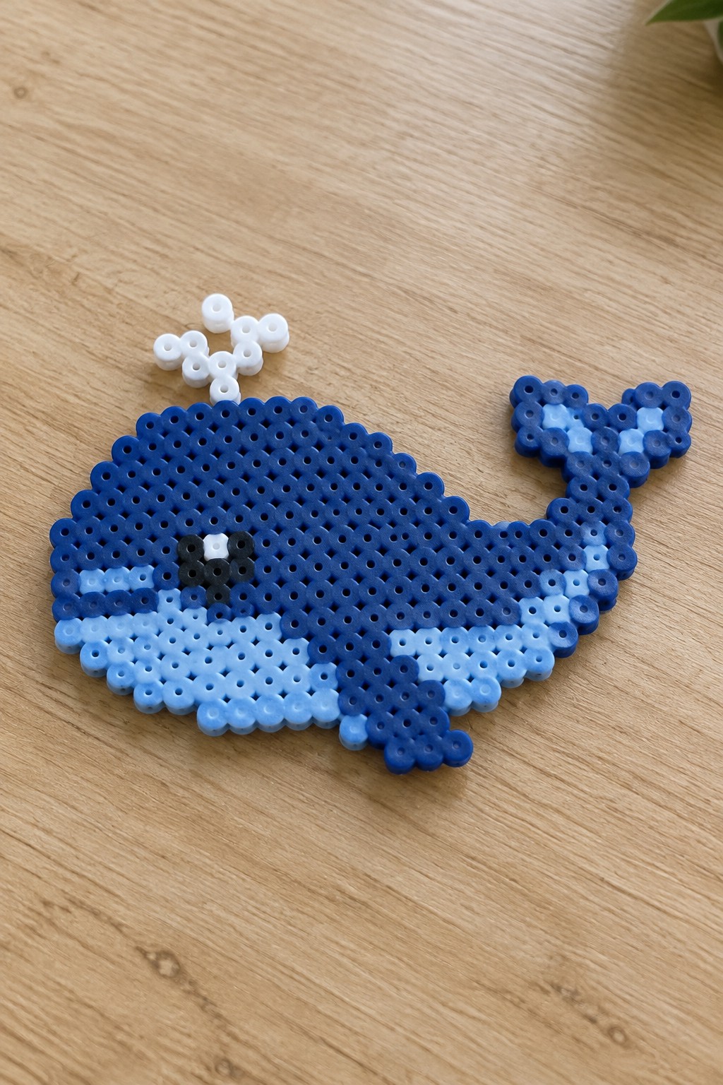 Blue perler bead whale with eye and white spout on wooden surface