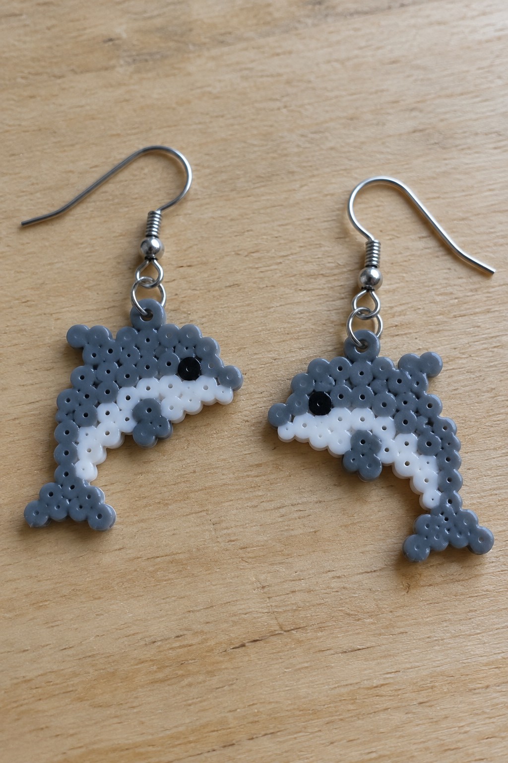 Pair of gray and white perler bead dolphin earrings with silver hooks on wood.