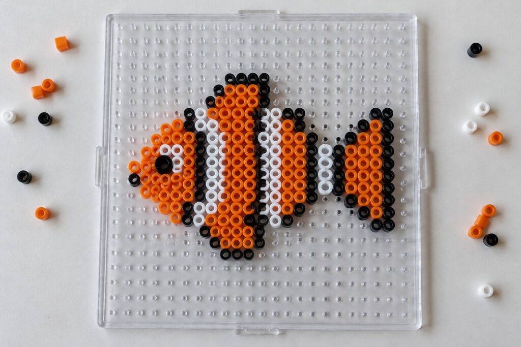 21 Ocean Perler Bead Ideas Featuring Aquatic Animals Clownfish Perler bead pattern in orange, white, and black on a pegboard.