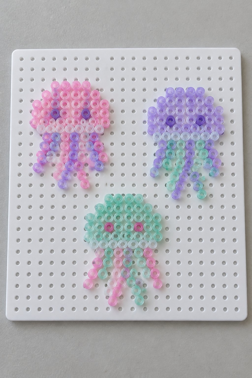 Three colorful perler bead jellyfish in pink, purple, and mint on white pegboard.