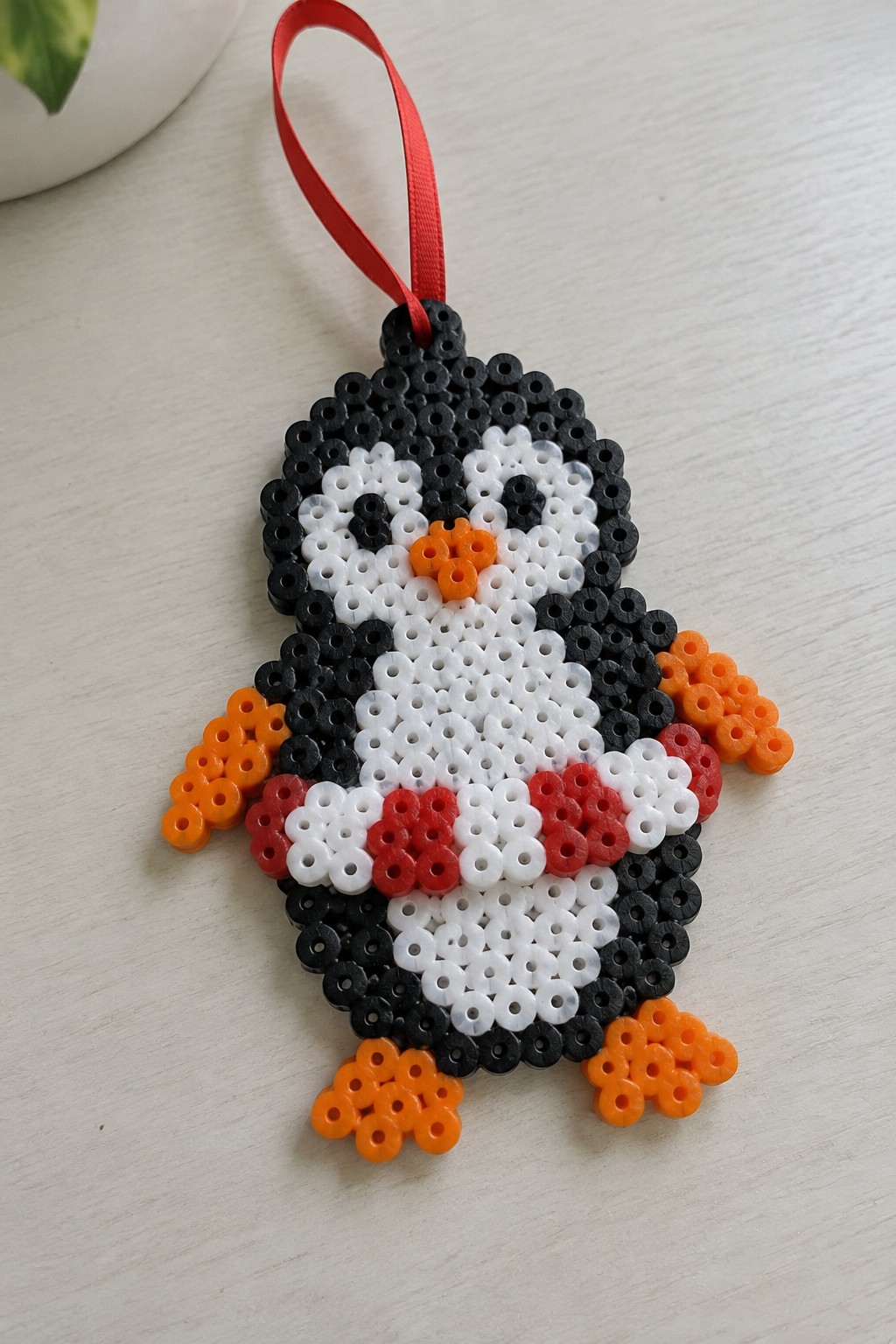 Perler bead penguin ornament with black body, orange feet, and red ribbon