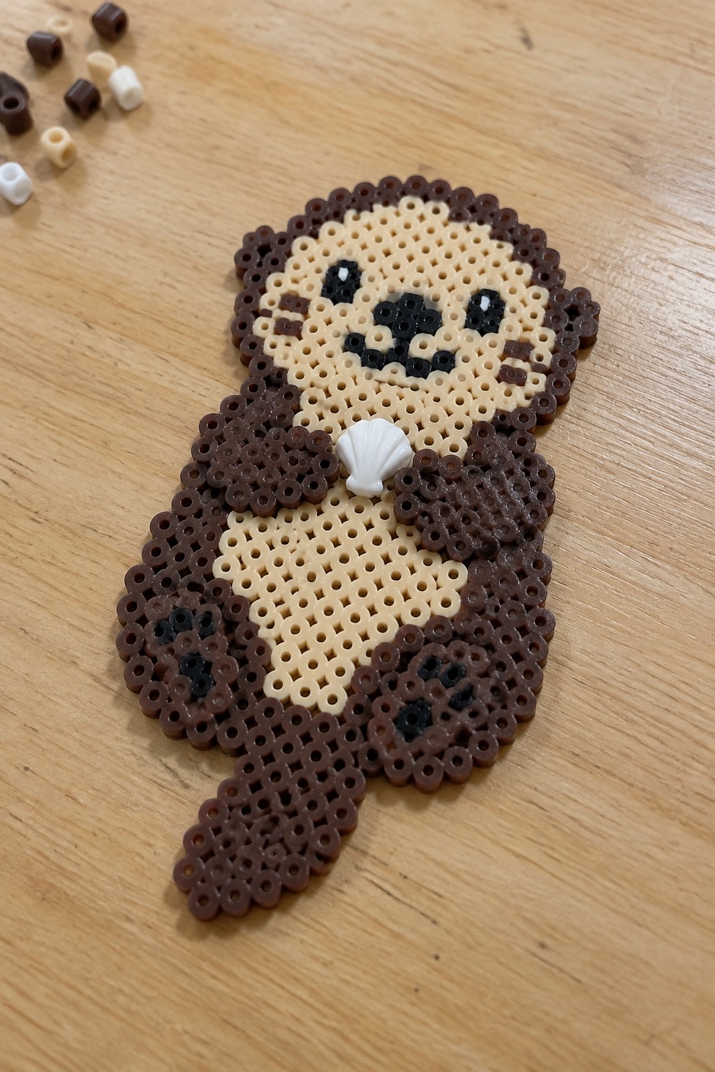Brown perler bead otter with beige belly holding white seashell on wood