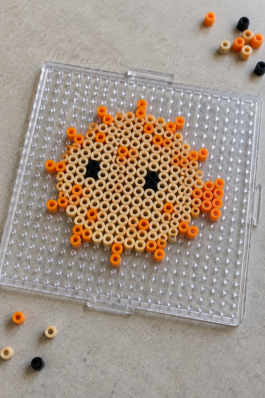 Perler beads forming a fox face on a clear plastic pegboard with loose beads nearby.