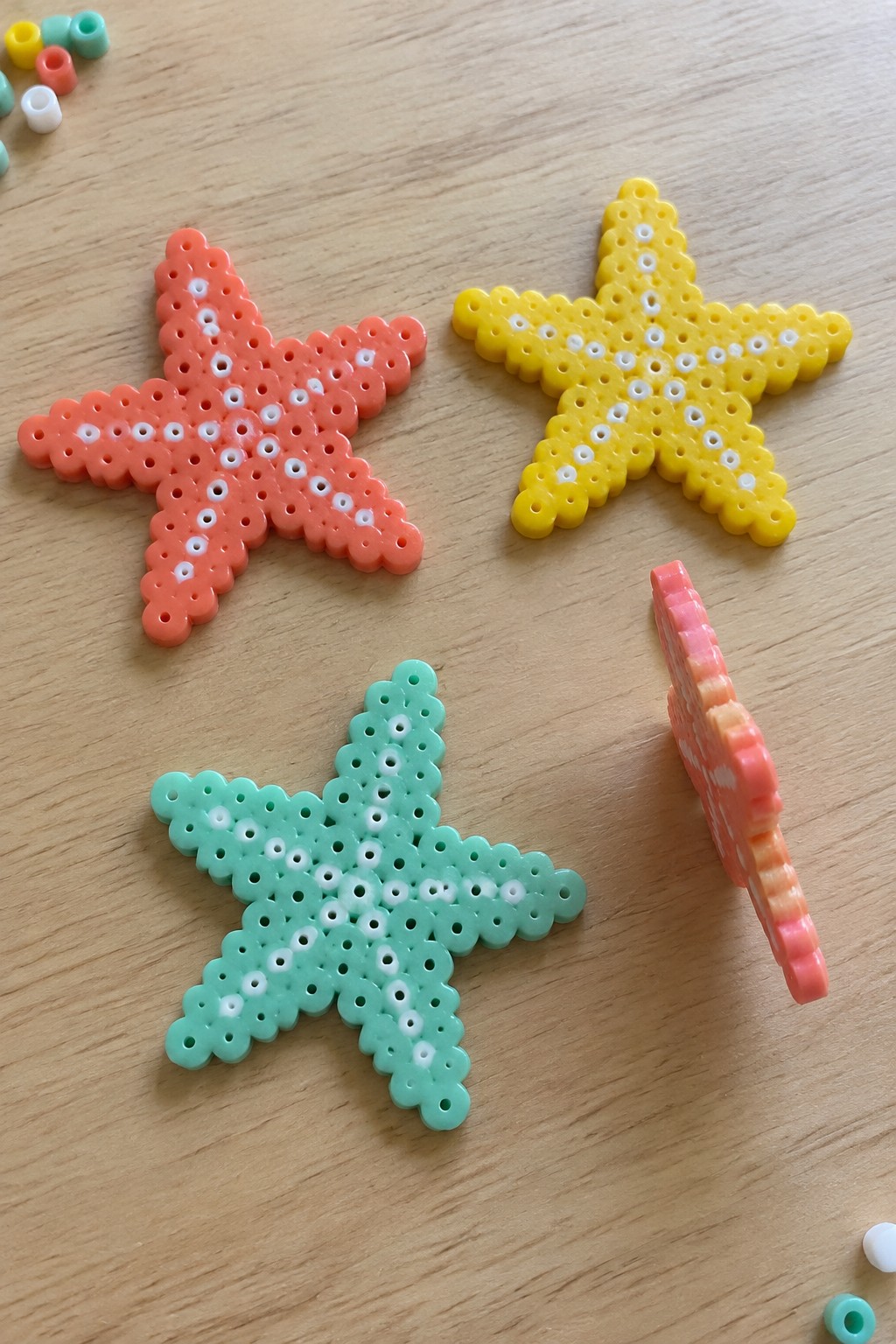 Three fused Perler bead starfish in orange, yellow, and mint green with white inner patterns, shown on a wooden table with a small side-view piece nearby.