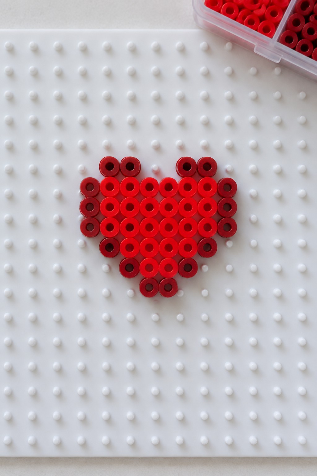 Red heart made of beads on white pegboard beside open red bead container.