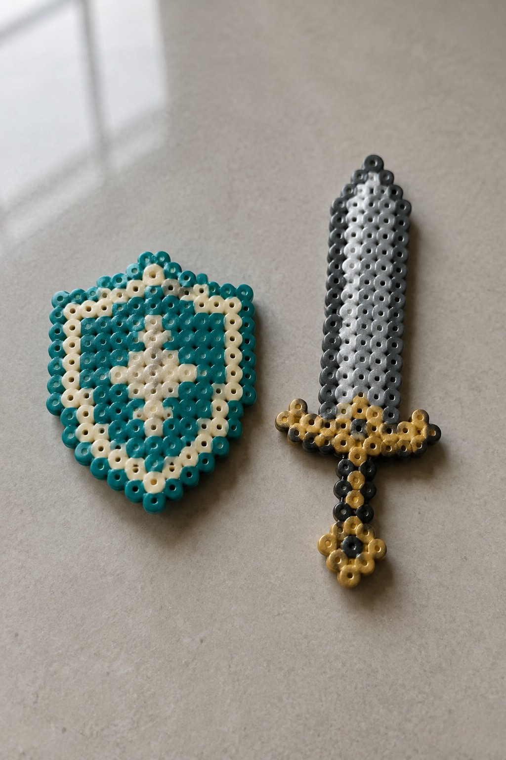 Teal and white Perler bead shield beside gray and gold beaded sword on table