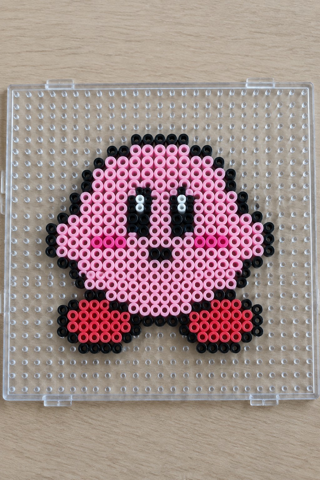 Kirby Perler bead sprite on a pegboard with pink body, black outline, red feet, and white eye highlights.