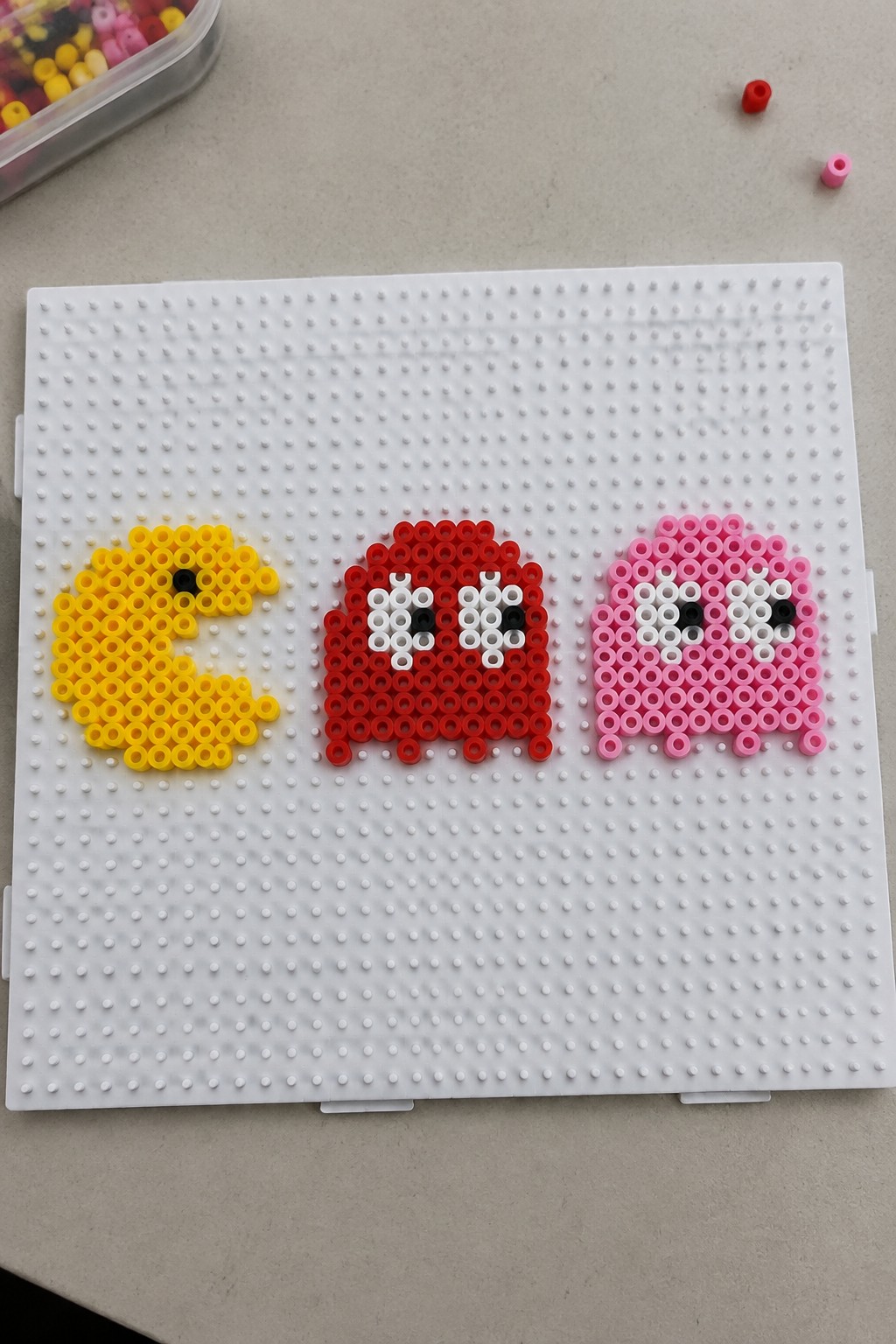 Yellow Pac-Man with red and pink ghost figures on white perler bead pegboard.