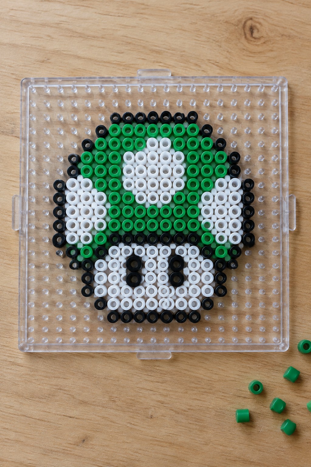 Clear pegboard with green, white, and black perler beads forming circular pattern on wood.