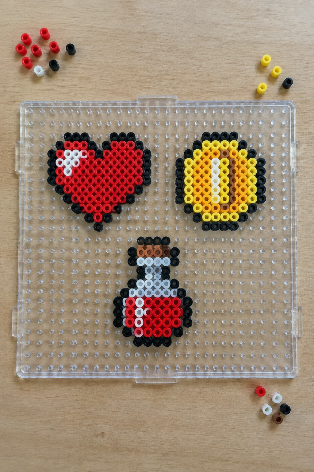 Three unfused Perler bead patterns on a pegboard showing a red heart sprite, a yellow circular sprite, and a small multi-colored character sprite.
