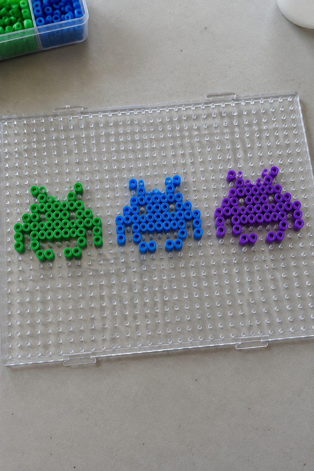 Green apple, blue fish, and purple car perler bead designs on clear pegboard.