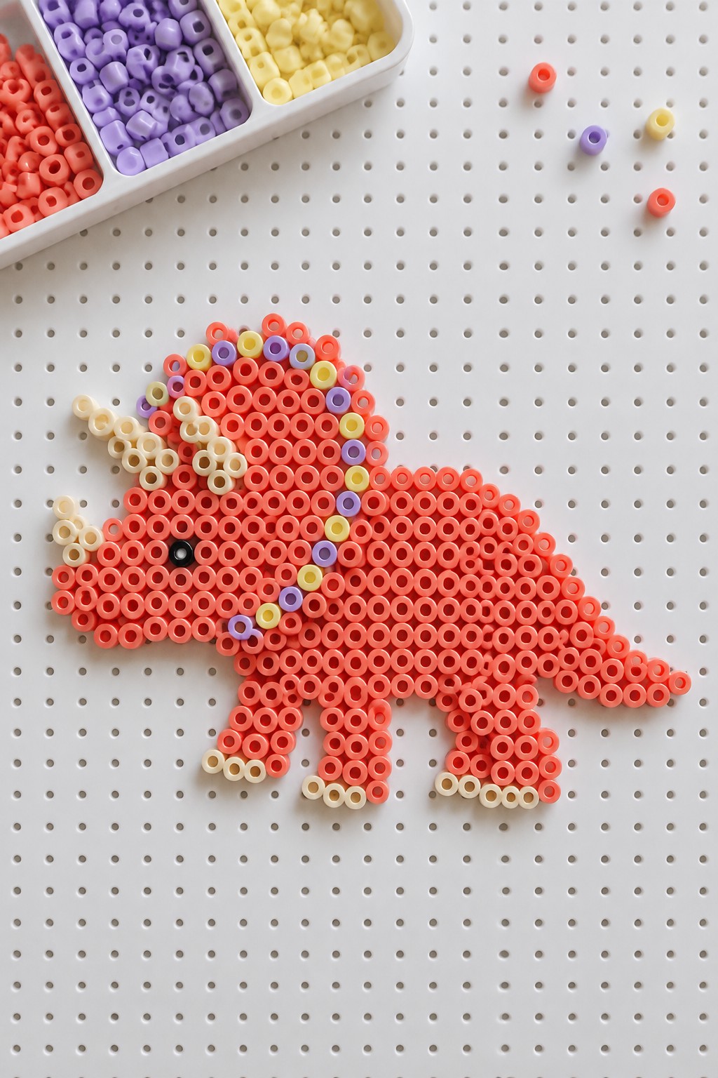 Pink perler bead triceratops with colorful accents on white pegboard beside bead tray
