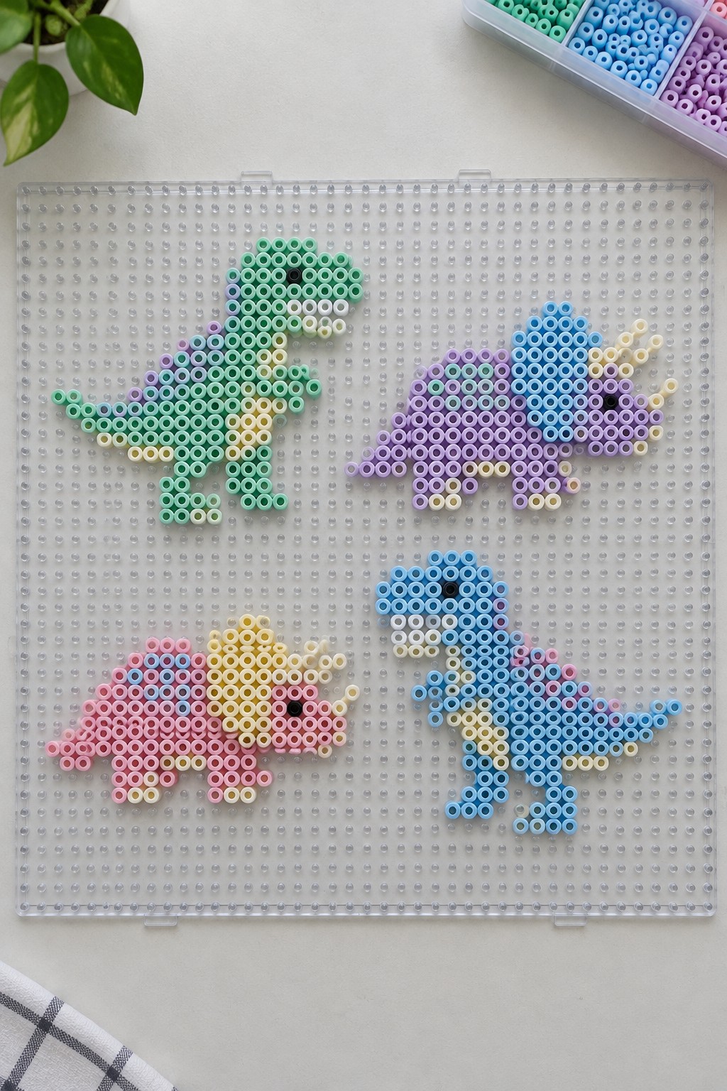 Four dinosaur shapes made from colorful Perler beads arranged on a clear pegboard.