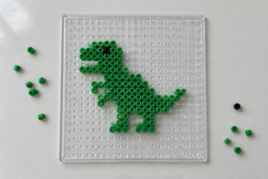 22 Dinosaur Perler Bead Patterns Featuring T-Rexes and Triceratops Green Perler beads form a T-Rex dinosaur on a clear pegboard with scattered loose beads.