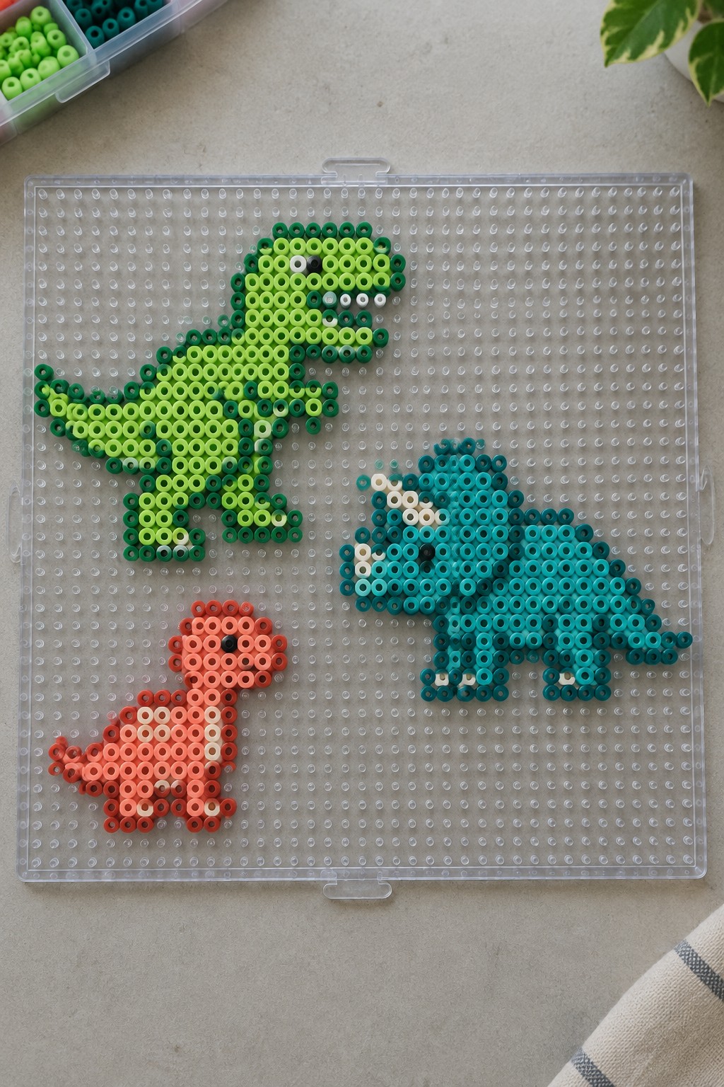 Three colorful perler bead dinosaurs on a clear plastic pegboard with beads nearby