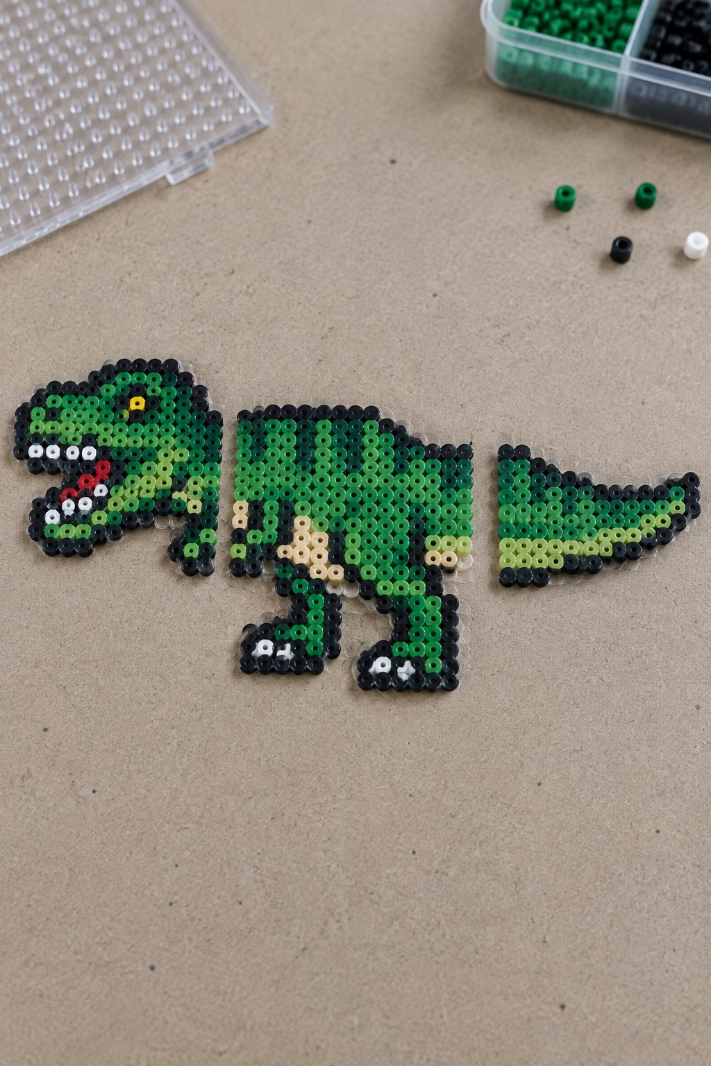 Perler bead T-Rex dinosaur arranged in three unfused sections on a pegboard using shades of green with black outlines.