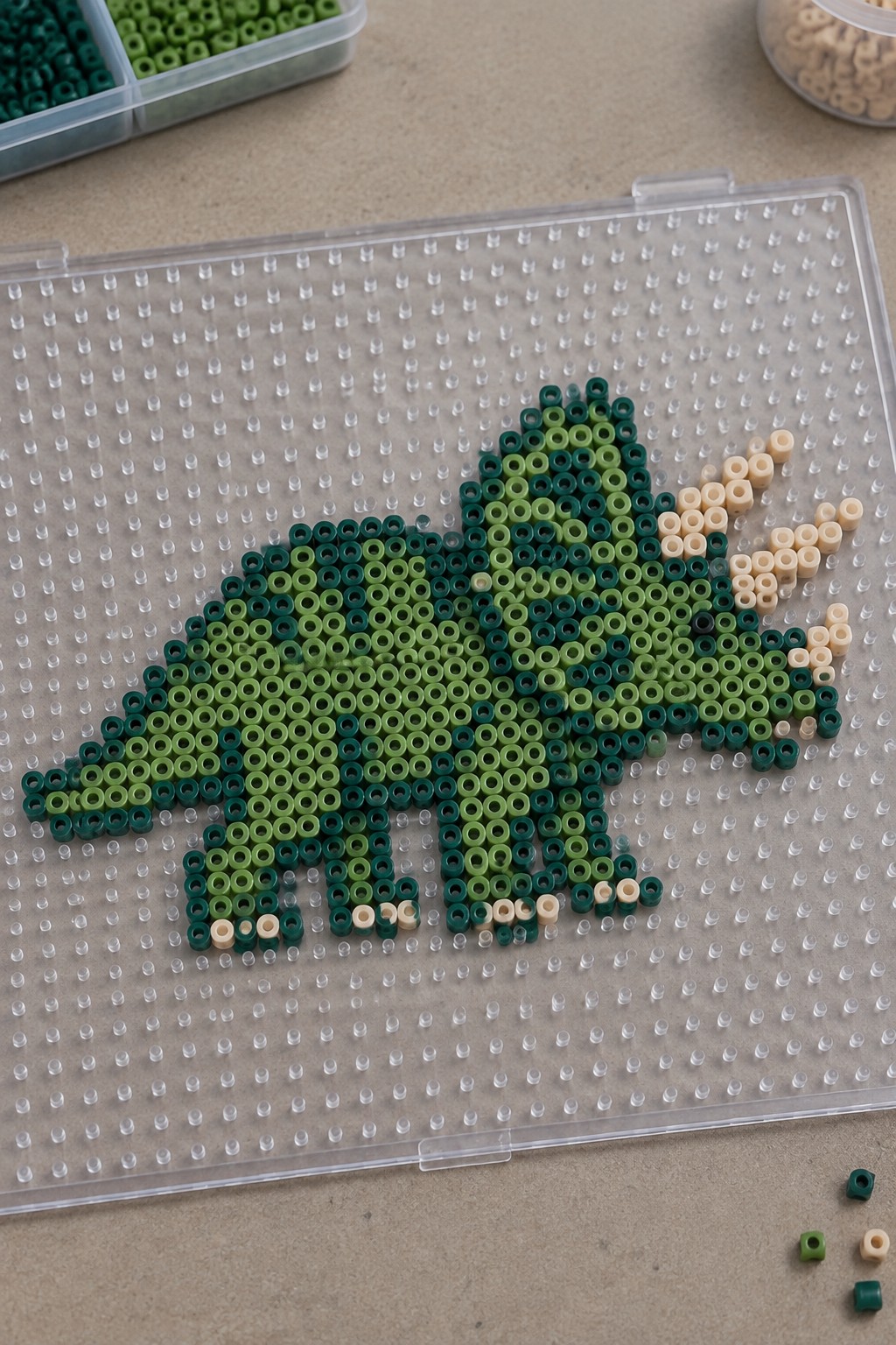 Gradient green perler bead dinosaur on clear pegboard with beige accents.