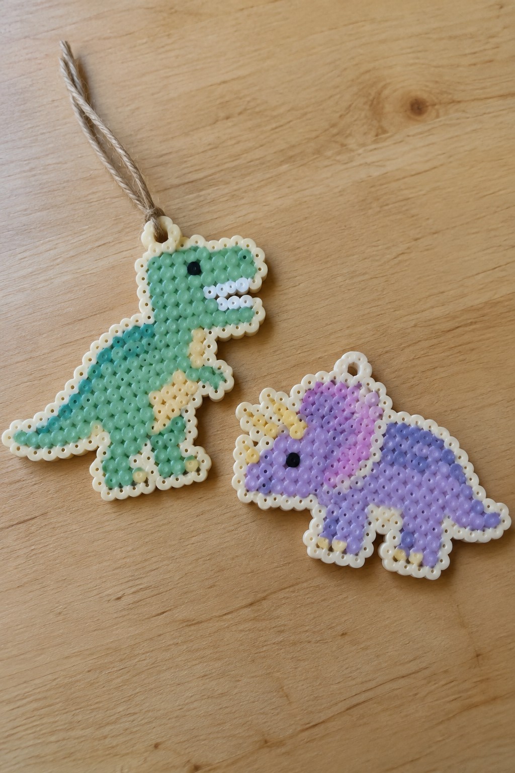 Two fused Perler bead dinosaur ornaments, a green T-Rex and a purple Triceratops with white borders, on a wooden surface.
