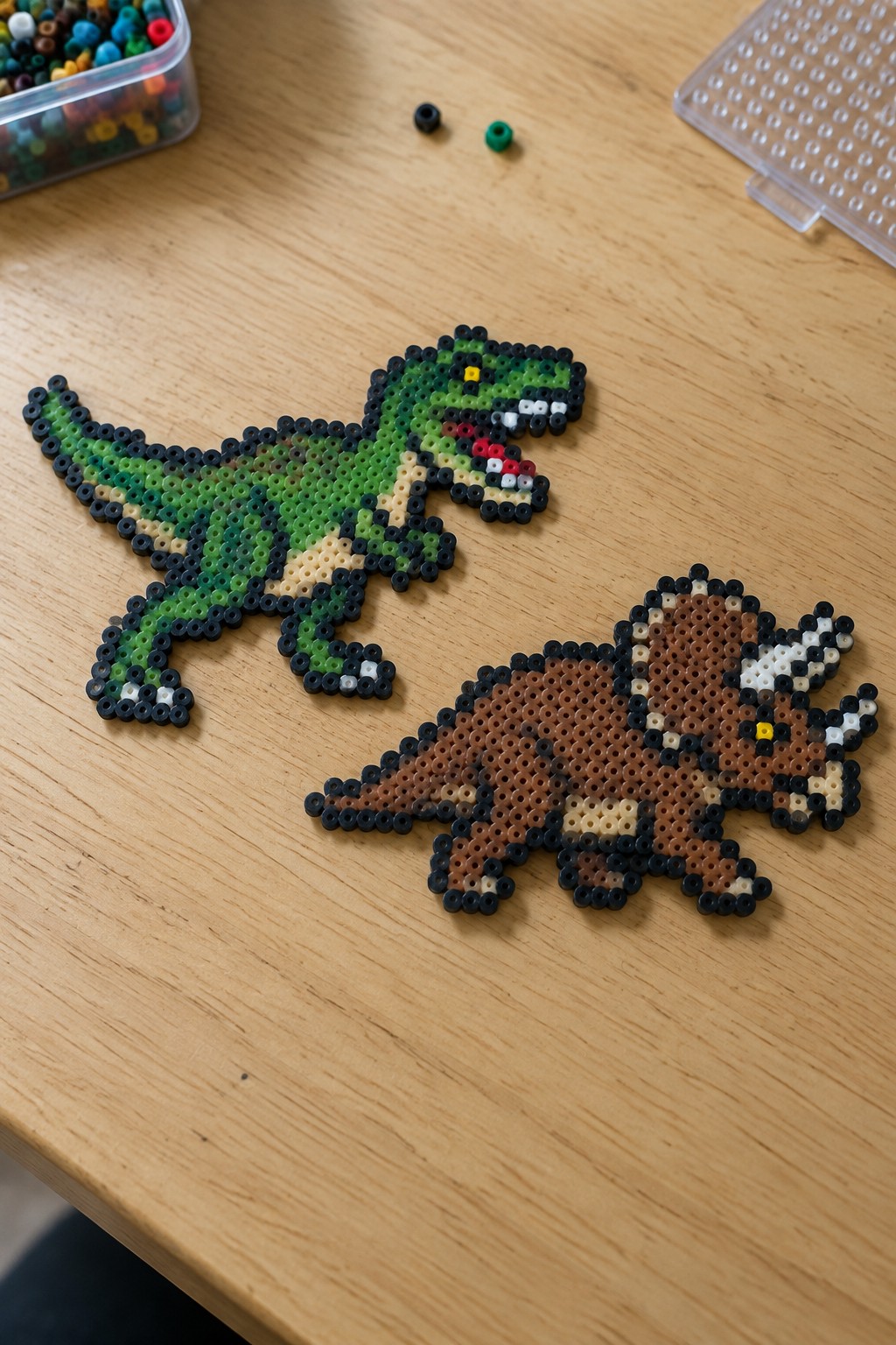 Two unfused Perler bead dinosaurs on a table: a green T-Rex and a brown Triceratops.
