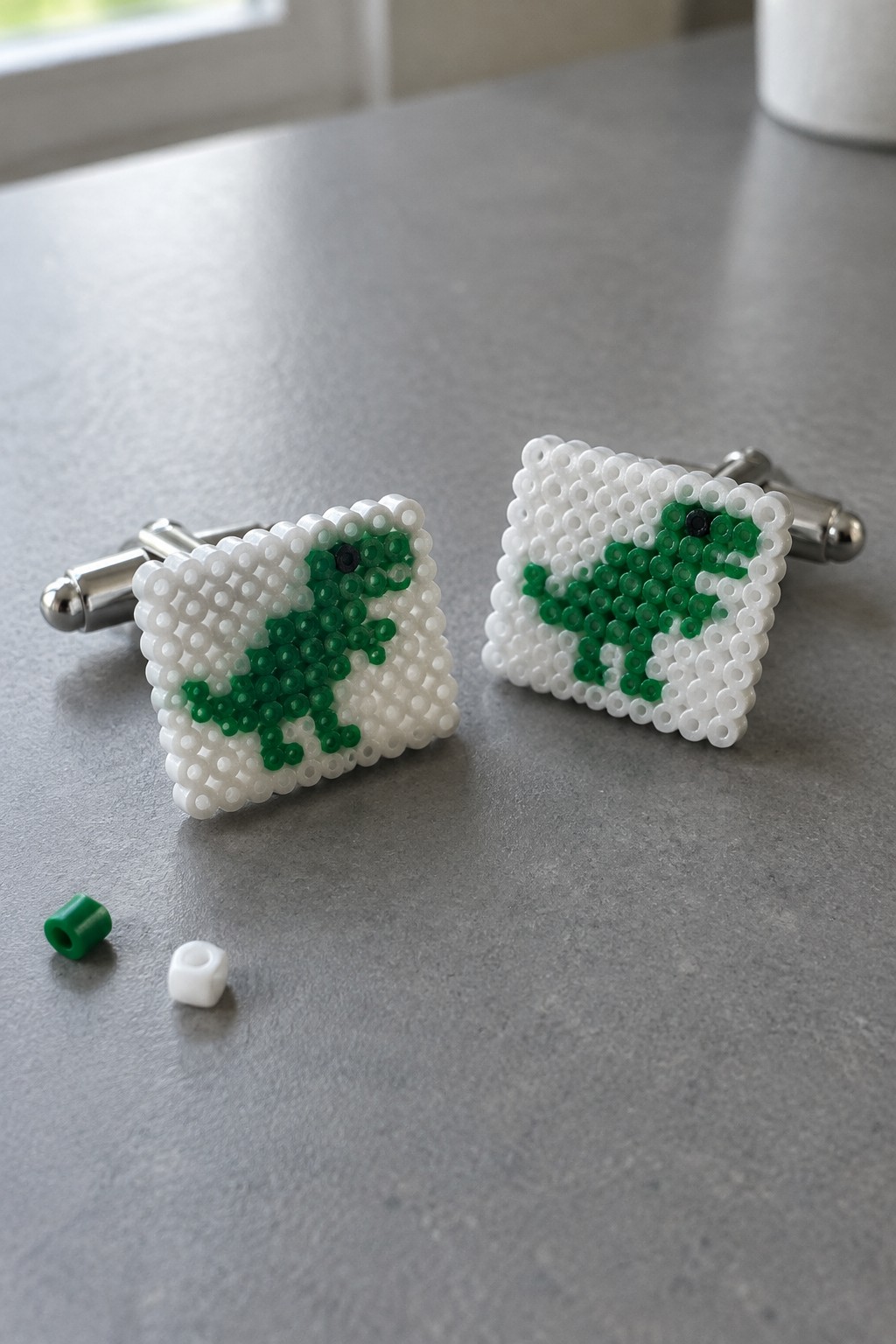 Two perler bead cufflinks with green dinosaur designs and metal backs on gray surface.
