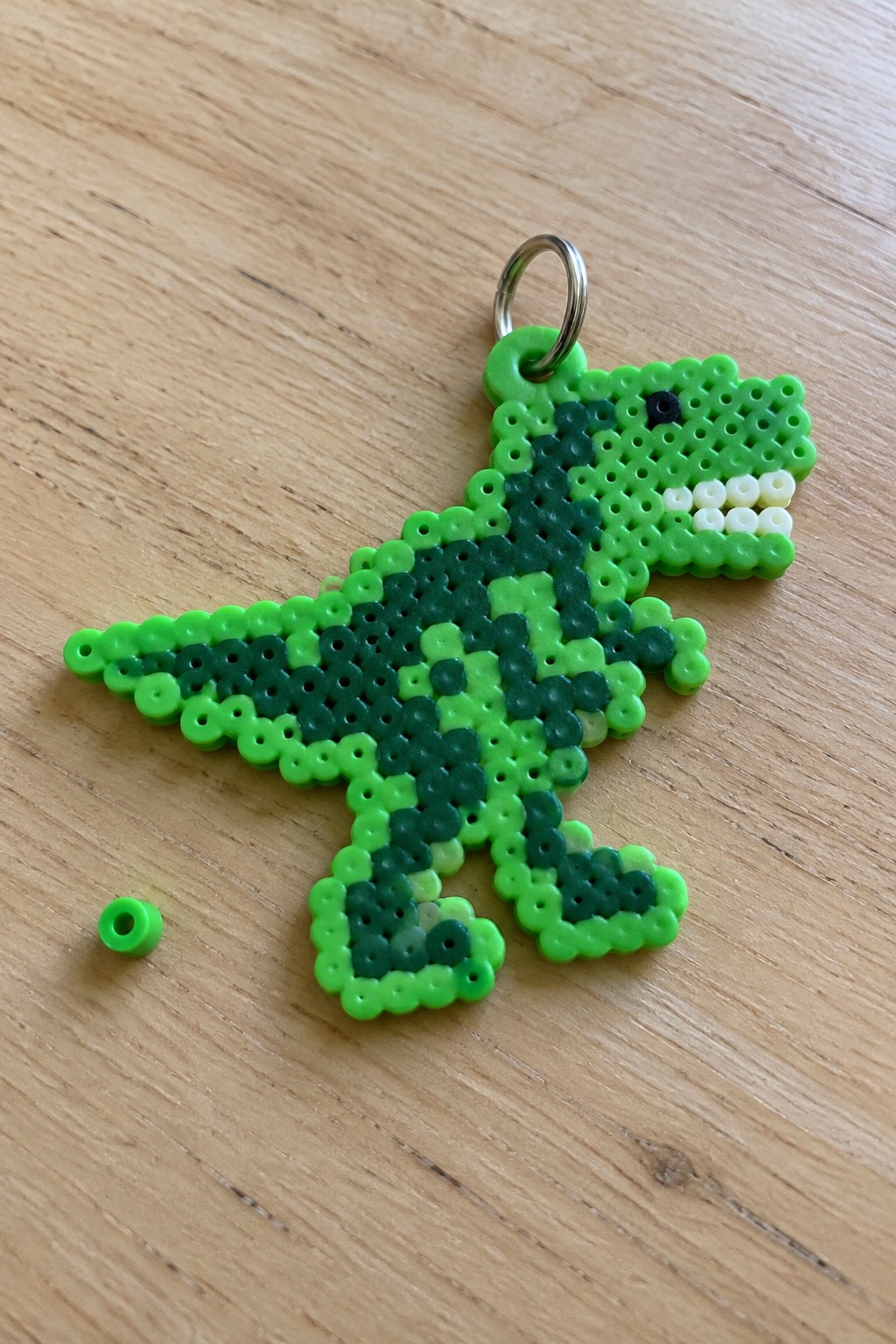 A fused Perler bead T-Rex keychain made from multiple shades of green beads with white teeth and a black eye, attached to a metal ring.