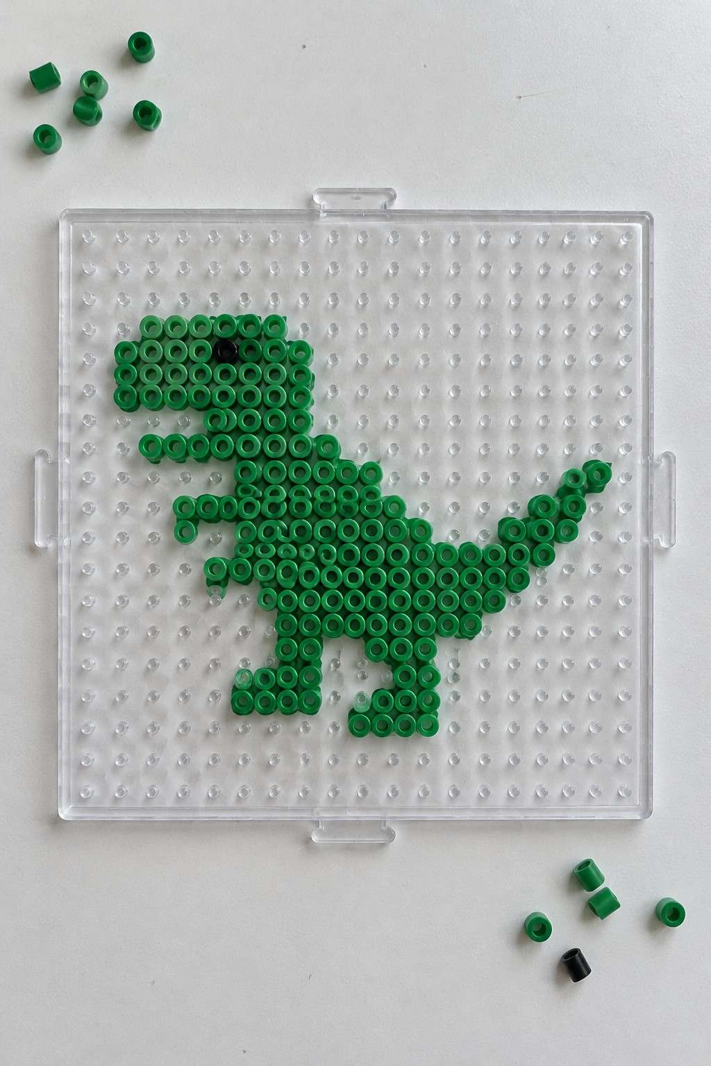 Green Perler beads form a T-Rex dinosaur on a clear pegboard with scattered loose beads.