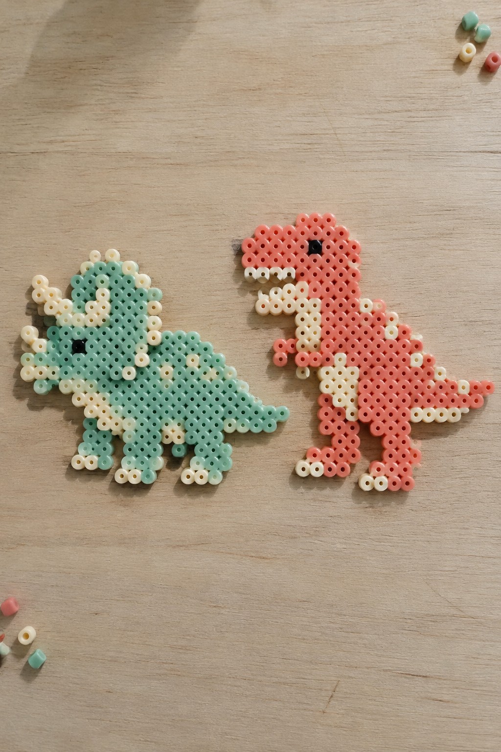 Mint green triceratops and pink T-rex perler bead dinosaurs on wood.