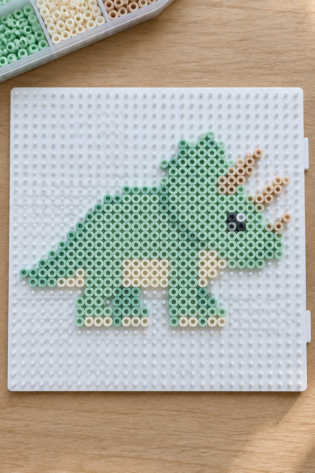 Green and beige Perler beads forming a dinosaur on a pegboard with bead tray.