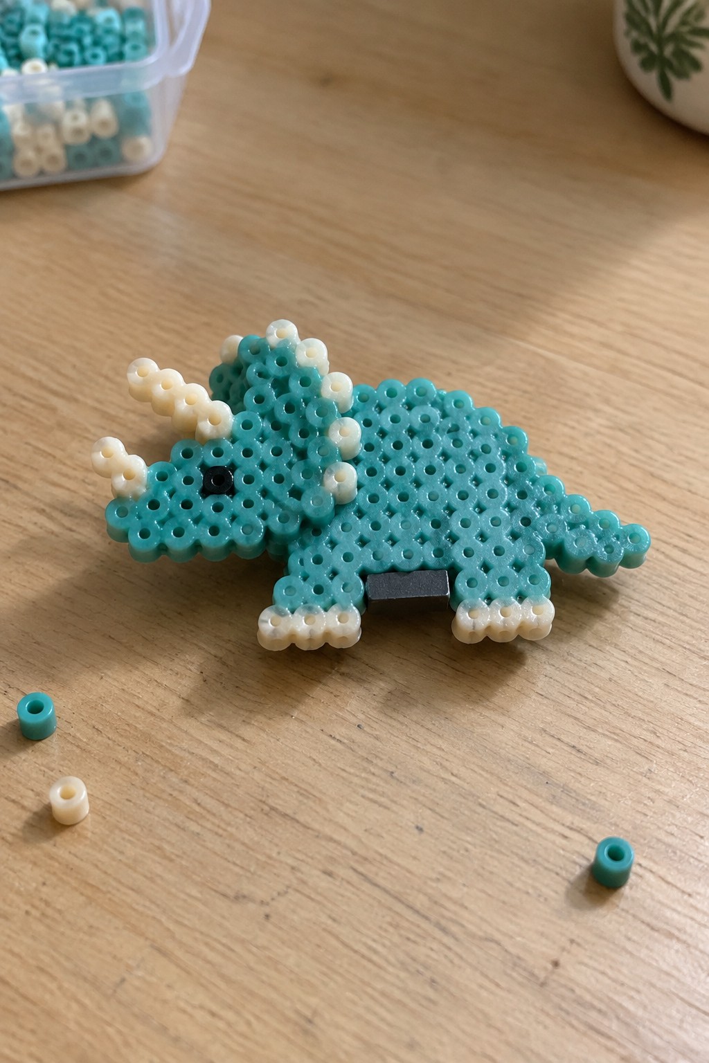 Turquoise and white perler bead triceratops dinosaur on wooden table with loose beads.