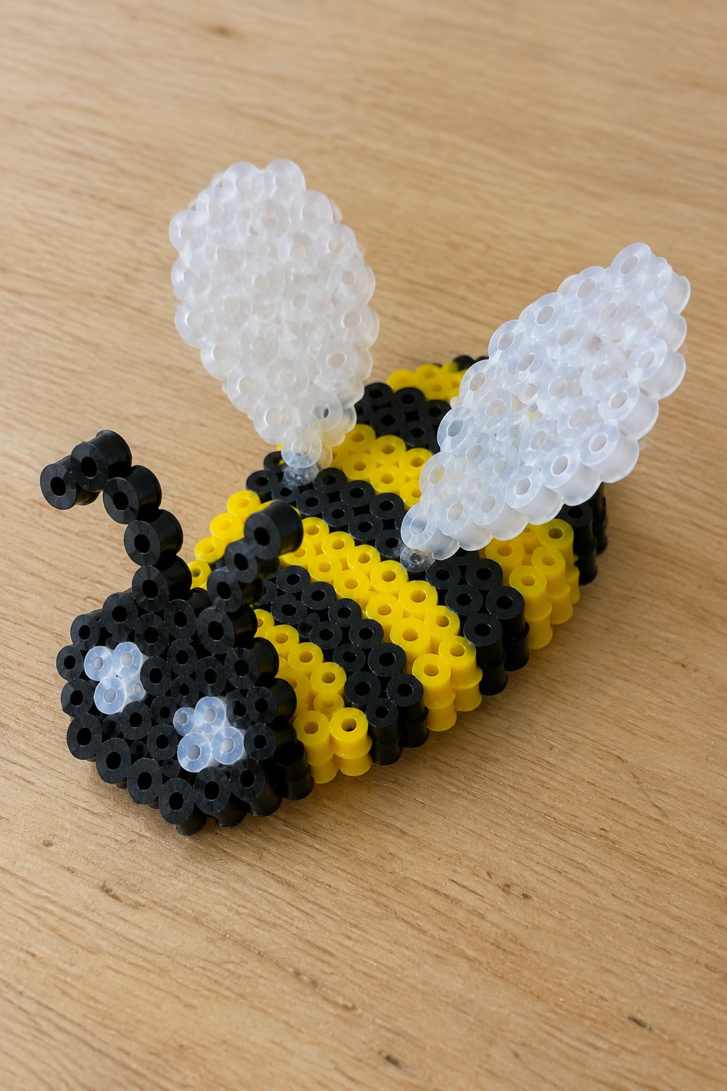 Yellow and black striped perler bead bee with white wings on wood.