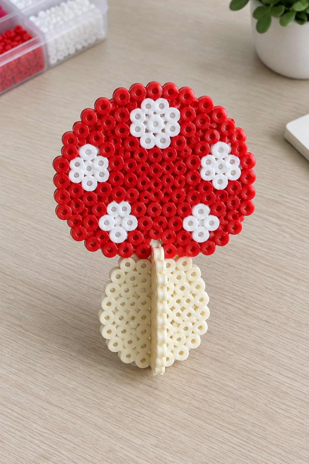 A fused Perler bead mushroom with a red cap, white flower-shaped spots, and beige stem standing upright.