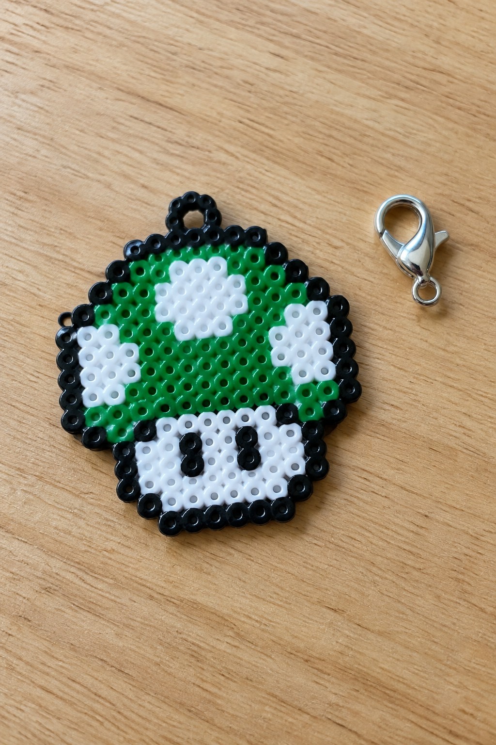 Green white black perler bead Mario mushroom charm beside silver metal clasp on wood
