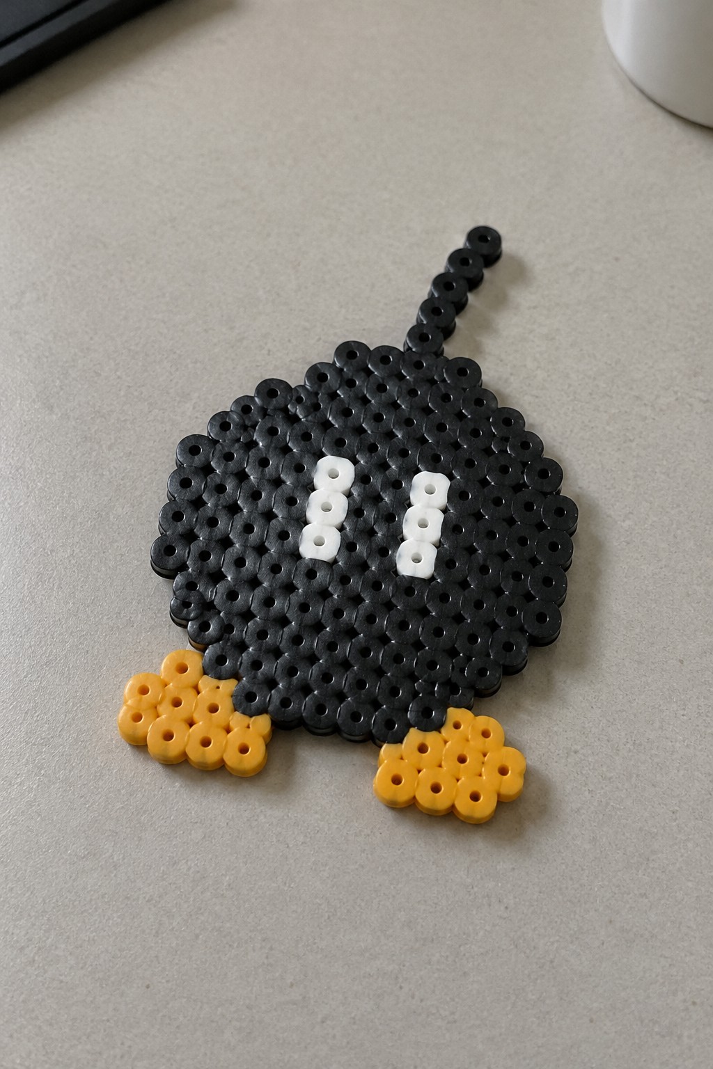 Perler bead Boo ghost on a pegboard with black body, white eyes, yellow feet, and black tail.