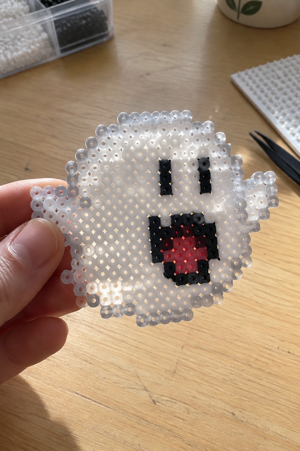 Hand holding white perler bead ghost with black eyes and red mouth on wood table