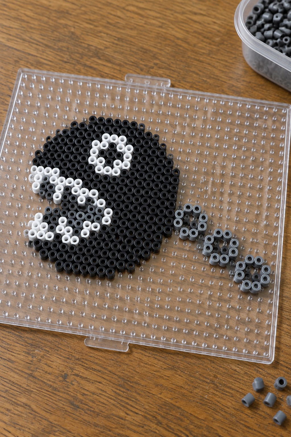 Clear pegboard with black and white Perler beads forming a design beside loose gray beads