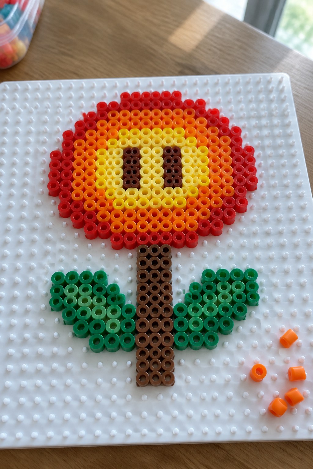 Perler bead flower with red-orange-yellow petals, brown stem, green leaves on pegboard.