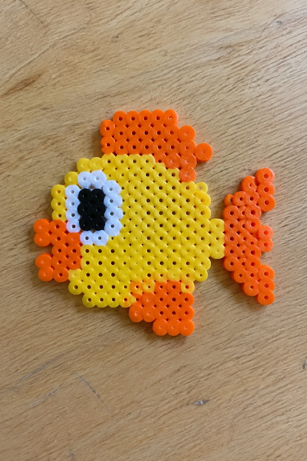 Perler bead fish on a pegboard with yellow body, orange fins and tail, and black-and-white eye.